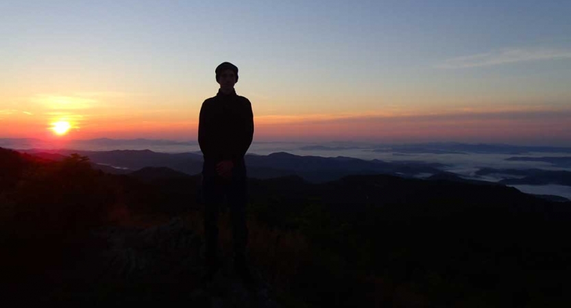 The silhouette of a person is illuminated by the sun behind them. The sky appears in soft shades of blue, yellow, orange, pink and purple.